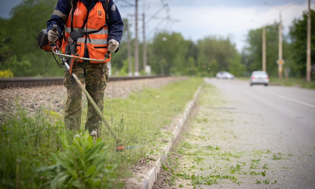 Worker in special protective reflective clothing with a lawn mower in his hands, mows grass with dandelions next to the road and railway tracks. Trimmer in the hands of a man. Cars in the background.