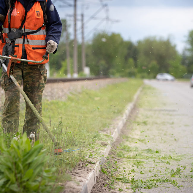 Worker in special protective reflective clothing with a lawn mower in his hands, mows grass with dandelions next to the road and railway tracks. Trimmer in the hands of a man. Cars in the background.