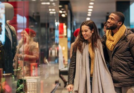A smiling interracial couple shop in a mall while wearing winter clothing.