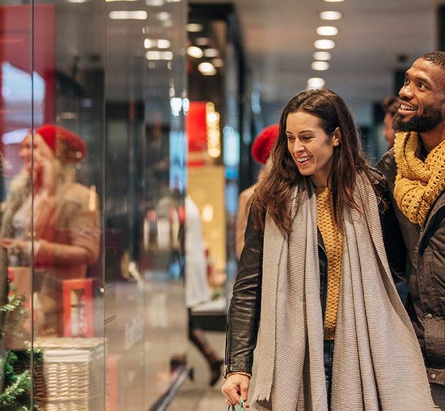 A smiling interracial couple shop in a mall while wearing winter clothing.